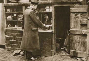 Un zapatero, establecido en un puesto improvisado en un callejón adoquinado de Mayfair, recibe botas de un chófer, de Maravilloso Londres, publicado 1926-27